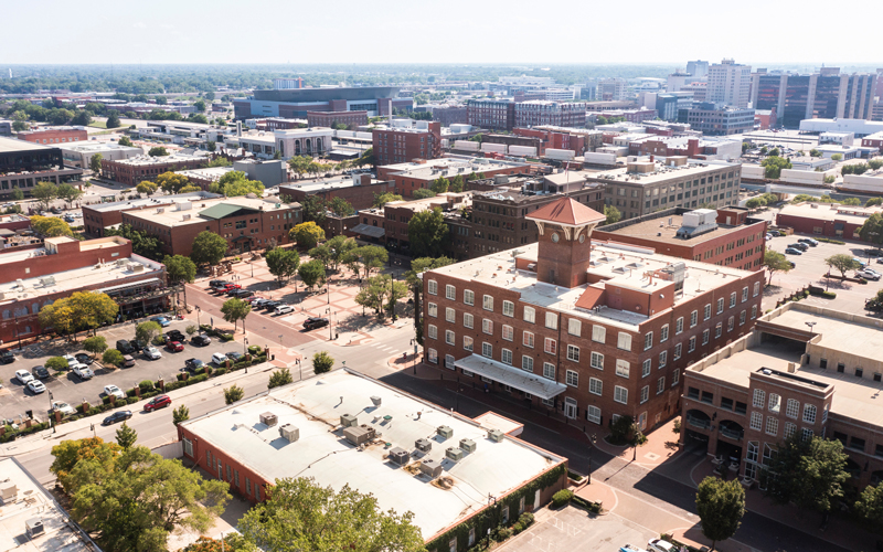 An aerial view of a mid-sized city captures a mix of historic and modern government buildings, tree-lined streets, parking areas, and civic infrastructure. At the center of the image is a prominent brick municipal building with a clocktower, symbolizing local governance and community leadership. This image represents the operational demands of cities and municipalities, where timely mailings—such as utility bills, tax notices, public announcements, and certified letters—are critical for citizen communication and compliance. Postalocity’s secure, scalable platform helps government offices streamline mail workflows, reduce costs, and maintain service reliability across departments.