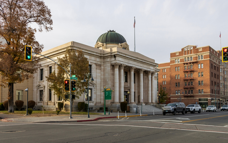 A stately government building with neoclassical architecture stands prominently at a street corner, featuring tall columns, a domed roof, and an American flag flying above. Traffic lights and parked cars indicate an active municipal setting, while adjacent buildings hint at a blend of history and civic activity. This image represents county offices responsible for vital communications such as tax statements, voting information, property records, and court notices. Postalocity’s mailing platform helps county departments automate and streamline these processes—ensuring timely, secure delivery while reducing manual workload and administrative costs.