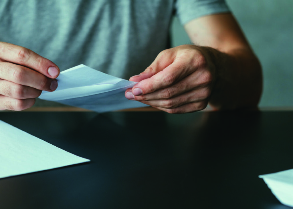 A close-up of a person folding a piece of paper and inserting it into a white envelope, seated at a dark table. The individual is engaged in the repetitive, time-consuming task of manual mailing—representing the traditional process of sending patient statements or healthcare notices. This image illustrates the inefficiencies that many medical offices still face. Postalocity eliminates these burdens by automating printing, folding, stuffing, and postage. For healthcare teams, that means more time focused on patients and less time spent on paperwork—improving both operational efficiency and patient experience.