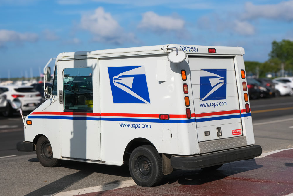 A USPS mail delivery truck is parked on a street near a public lot, clearly displaying the official red, white, and blue postal service branding and www.usps.com web address. The vehicle represents dependable, domestic mail distribution throughout the United States. For international organizations, this image symbolizes Postalocity’s advantage in bypassing cross-border mail delays by processing and mailing documents directly within the U.S., ensuring faster, more reliable delivery of time-sensitive materials such as contracts, compliance documents, or partner communications.