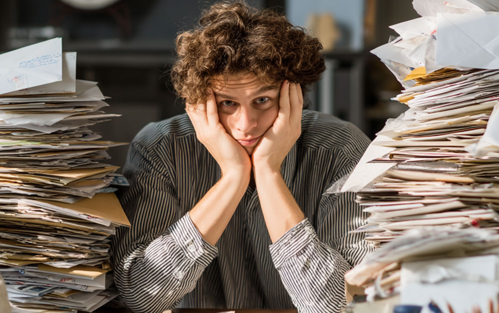 A visibly overwhelmed person sits at a desk with their face resting in their hands, surrounded by towering stacks of unsorted envelopes and paper mail. The cluttered scene captures the frustration and inefficiency of manual mailing processes often faced by city and municipal staff. This image underscores the burden of printing, folding, and stuffing thousands of documents for public notices, utility bills, and tax statements. Postalocity offers a powerful alternative—automating the entire mailing process so municipal teams can save time, reduce labor, and refocus efforts on serving their communities more effectively.