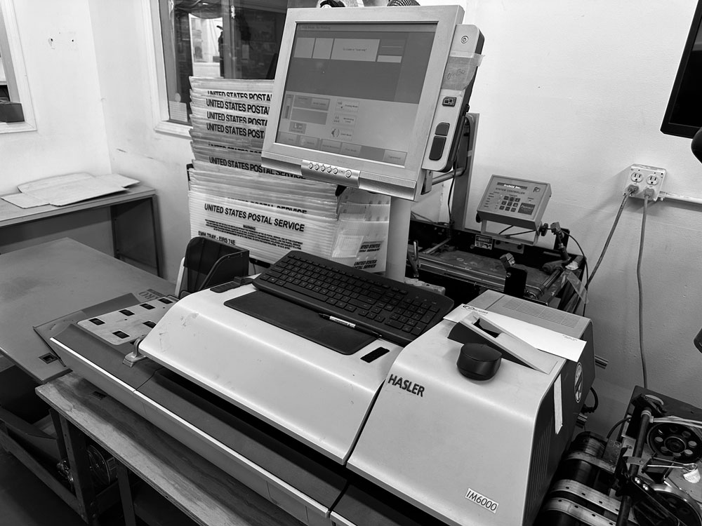 A black-and-white image of an outdated office postage meter and mailing station, including a Hasler IM6000 postage machine, keyboard, and monitor setup surrounded by trays labeled “United States Postal Service.” The workspace appears cluttered and labor-intensive, highlighting the burden of traditional mailing methods. This scene represents the kind of legacy equipment many county offices still rely on. Postalocity eliminates the need for such expensive hardware by offering a modern, cloud-based alternative with pay-as-you-go pricing—helping counties cut overhead costs, streamline mail operations, and free up resources for public service.