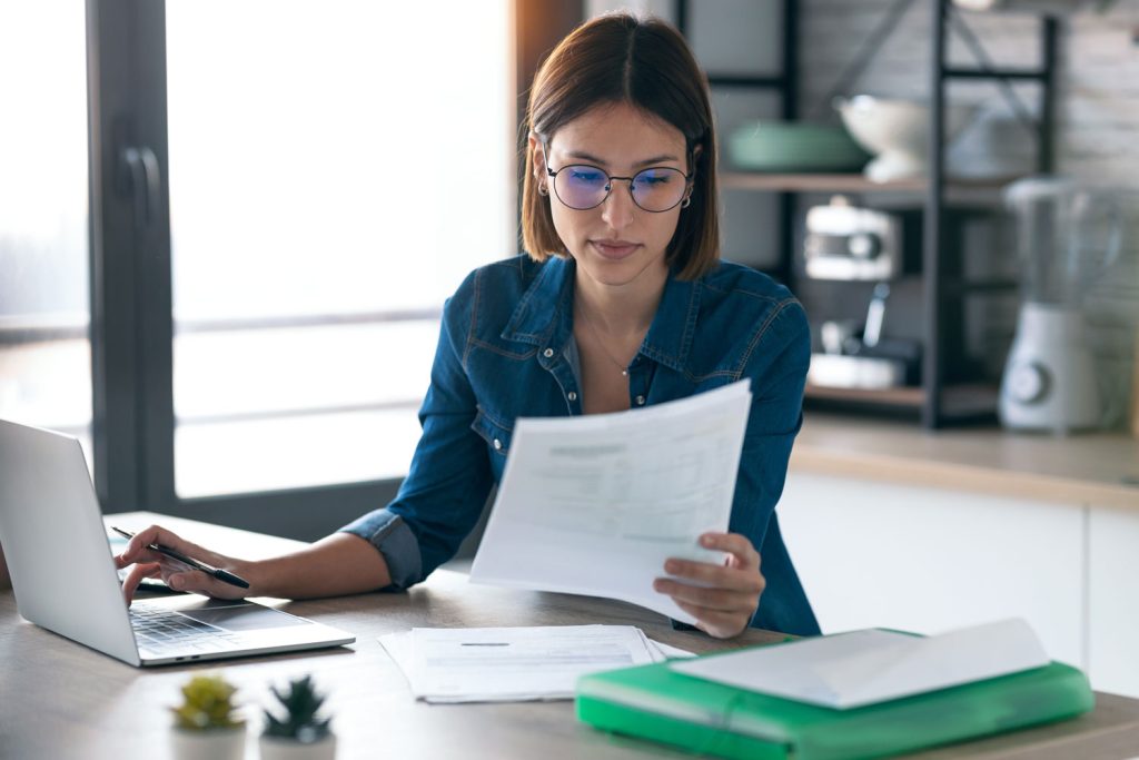 A woman with glasses sits at a kitchen table, attentively reviewing a printed invoice or billing statement while working on a laptop. Surrounded by papers and a green folder, she appears focused and organized, indicating a home-based or remote work setting. This image represents a small business owner or office administrator streamlining billing tasks—perfectly aligning with Postalocity’s efficient, secure business invoice mailing service that eliminates the need for manual processing and ensures timely delivery to clients or municipalities.