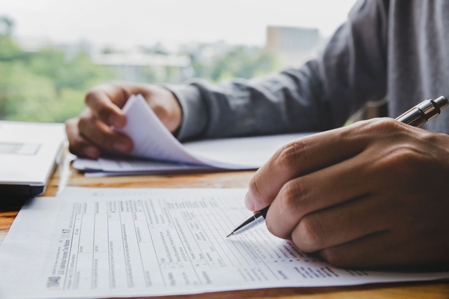 A close-up view of a person filling out an official IRS 1040 tax form with a pen, while flipping through a stack of paperwork. The individual sits at a desk near a window, with a laptop nearby, suggesting a work-from-home or small business setting. The image conveys attention to detail and urgency—ideal for illustrating the importance of accurate single mailings for sensitive documents like tax forms, legal contracts, or compliance notices. This visual supports Postalocity’s offering for single mailings with tools like PS Form 3817 for verified delivery.