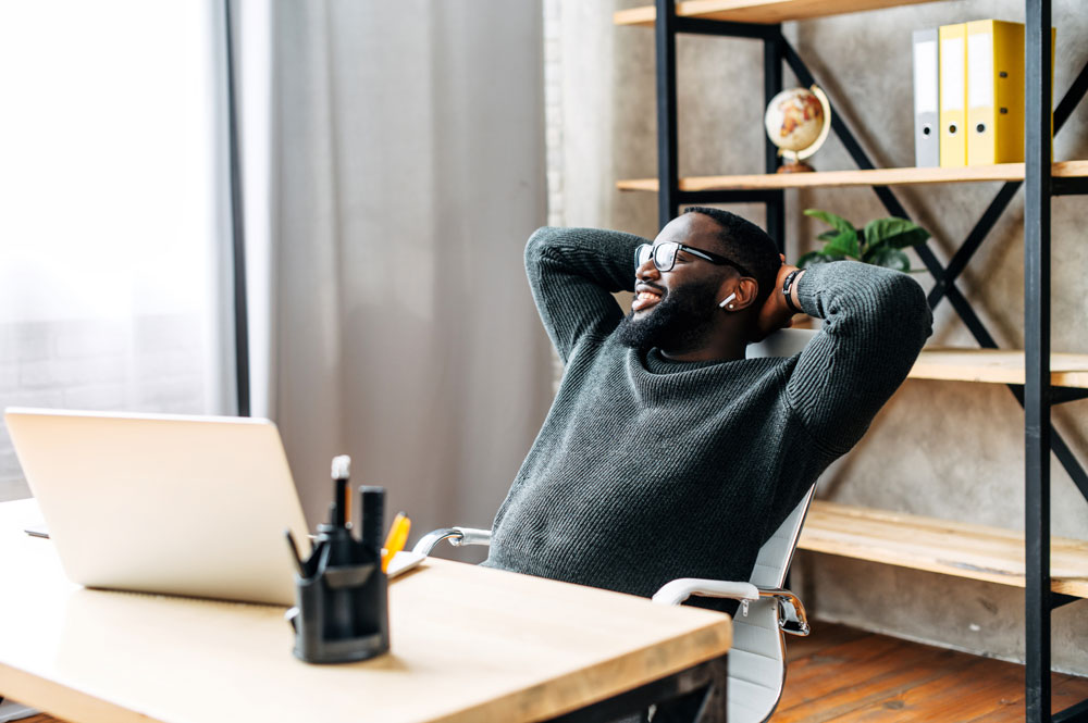A relaxed, smiling man leans back in his office chair with his hands behind his head, enjoying a moment of calm confidence. He wears a dark sweater and glasses, seated at a modern wooden desk with a laptop, office supplies, and shelves in the background. His posture reflects a sense of ease and relief, visually capturing the peace of mind that Postalocity delivers. The image illustrates the impact of Postalocity’s automated, secure mailing processes—with features like built-in checks and address verification—that reduce errors and free up time, allowing users to focus on what matters most, stress-free.