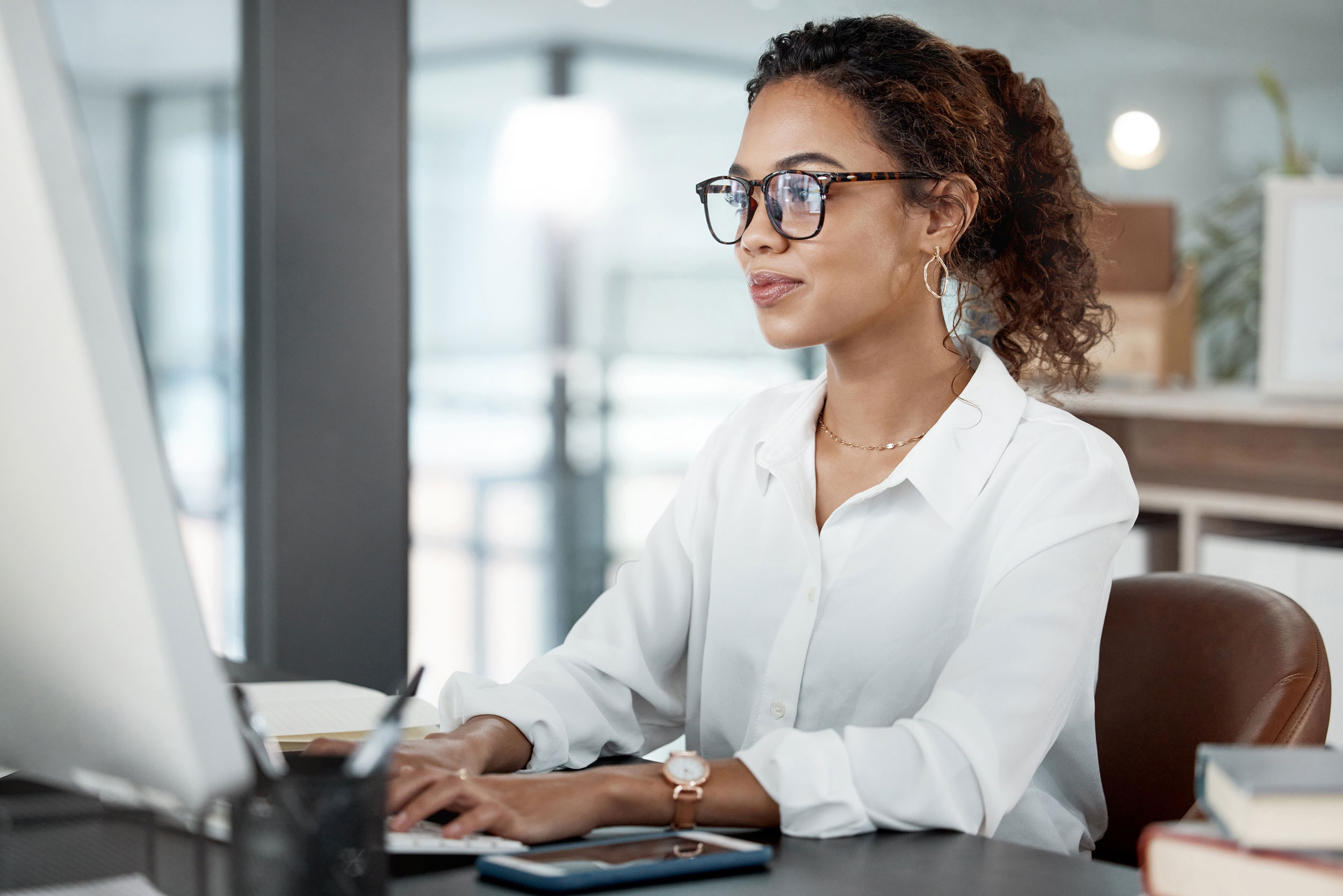 A professional woman sits at a sleek, modern desk, typing on a desktop computer in a bright, contemporary office environment. She wears a white blouse, tortoiseshell glasses, gold hoop earrings, and a rose-gold wristwatch, projecting a focused and polished demeanor. Books and a smartphone rest on the desk beside her, adding to the workspace's organized feel. Her expression and posture reflect concentration and confidence, representing someone managing official correspondence or business documents efficiently. The image illustrates the benefits of Postalocity’s cloud-based platform—making it easy to send business letters or legal-sized documents through USPS with speed, automation, and reliability.