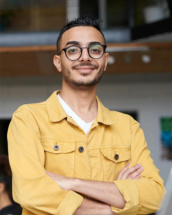 A confident young professional stands with arms crossed, smiling slightly in a bright, modern office environment. He is wearing round eyeglasses and a mustard yellow utility-style jacket over a white V-neck shirt. This image represents John D., City Clerk for Dodge City, Kansas, who praises Postalocity for transforming the way his office handles municipal mailings. According to his testimonial, tasks that used to be stressful and time-consuming—like billing week—are now streamlined, punctual, and fully trackable, allowing his team to better serve the community. The improvement has saved them 40 staff hours each month. The image underscores Postalocity’s effectiveness for government offices and high-performing teams.