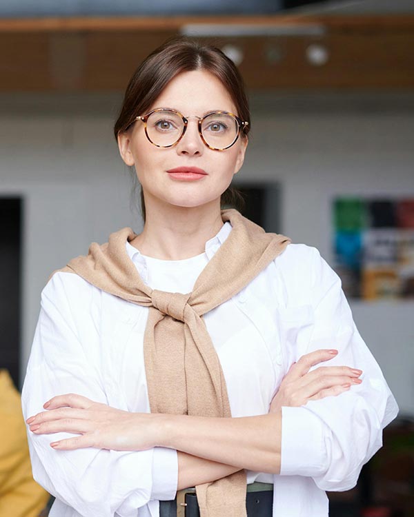 A composed and confident woman stands with her arms crossed in a contemporary office setting, wearing a white blouse with a tan sweater draped over her shoulders and tied loosely in front. She has round tortoiseshell glasses and a poised expression, reflecting leadership and assurance. This image represents Janet D., Client Services Lead at Global Services Ltd., who shares how Postalocity revolutionized her company’s U.S. mailing operations. By eliminating complexity and speeding up processes, Postalocity gave them the equivalent of a local office—without needing a physical one. Her team now saves $2,000 per month on postage processing alone, emphasizing the platform’s impact on international and remote business efficiency.