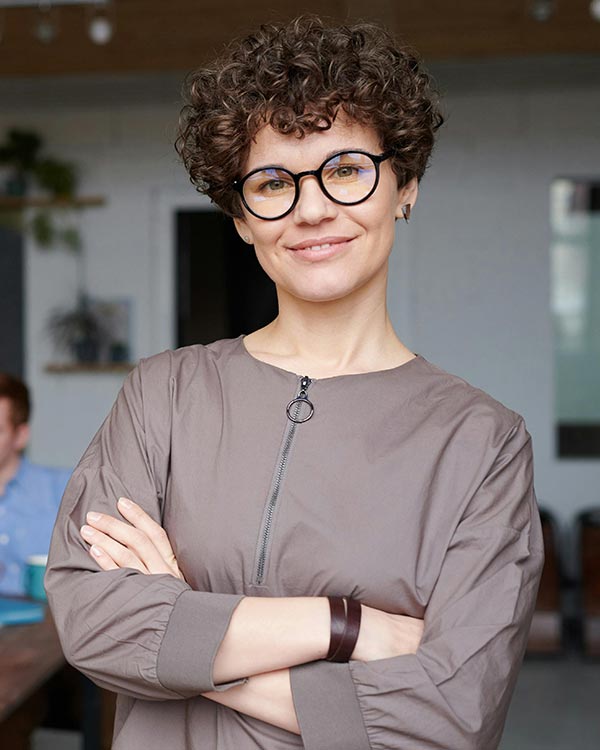 A confident professional woman stands smiling with arms crossed in a modern office setting, embodying the satisfaction and efficiency gained from using Postalocity. She wears a taupe zippered blouse, round eyeglasses, and a dark wrist cuff, conveying a smart, approachable demeanor. This image represents Jane D., the Operations Director at Heartland Credit Union, who praises Postalocity for transforming their mailing workflow. According to Jane, tasks that once took two full days now take less than an hour, allowing her team to prioritize member service over mailing logistics—an 80% reduction in prep time. The visual reinforces the platform’s value for high-performing teams across industries.
