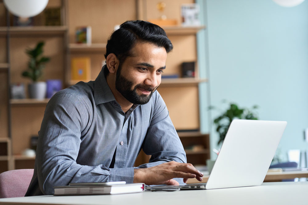 A focused, smiling man sits at a desk using a laptop, appearing relaxed and productive in a modern, well-lit workspace. He wears a button-up shirt and rests one hand on a notebook beside him, suggesting he’s working efficiently without the need for paper clutter. The image conveys the ease and simplicity of using Postalocity—where users can send a few or thousands of mail pieces with the same low-effort process. His calm demeanor reflects the platform’s promise: no stress, no complicated setup, no hidden fees—just a scalable, seamless mailing solution that grows with your business needs.